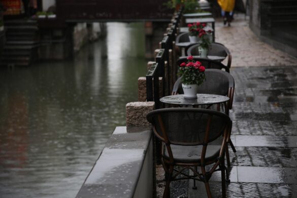 Verlassene Café-Terrasse an einem regnerischen Tag – nasse Stühle, leere Tische, rote Blumen in Blumentöpfen, spiegelnder Asphalt, melancholische Stimmung.