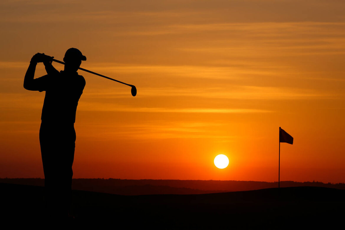 Silhouette eines Golfspielers bei Sonnenuntergang, mit Blick über das Fairway und einer Fahne am Horizont.