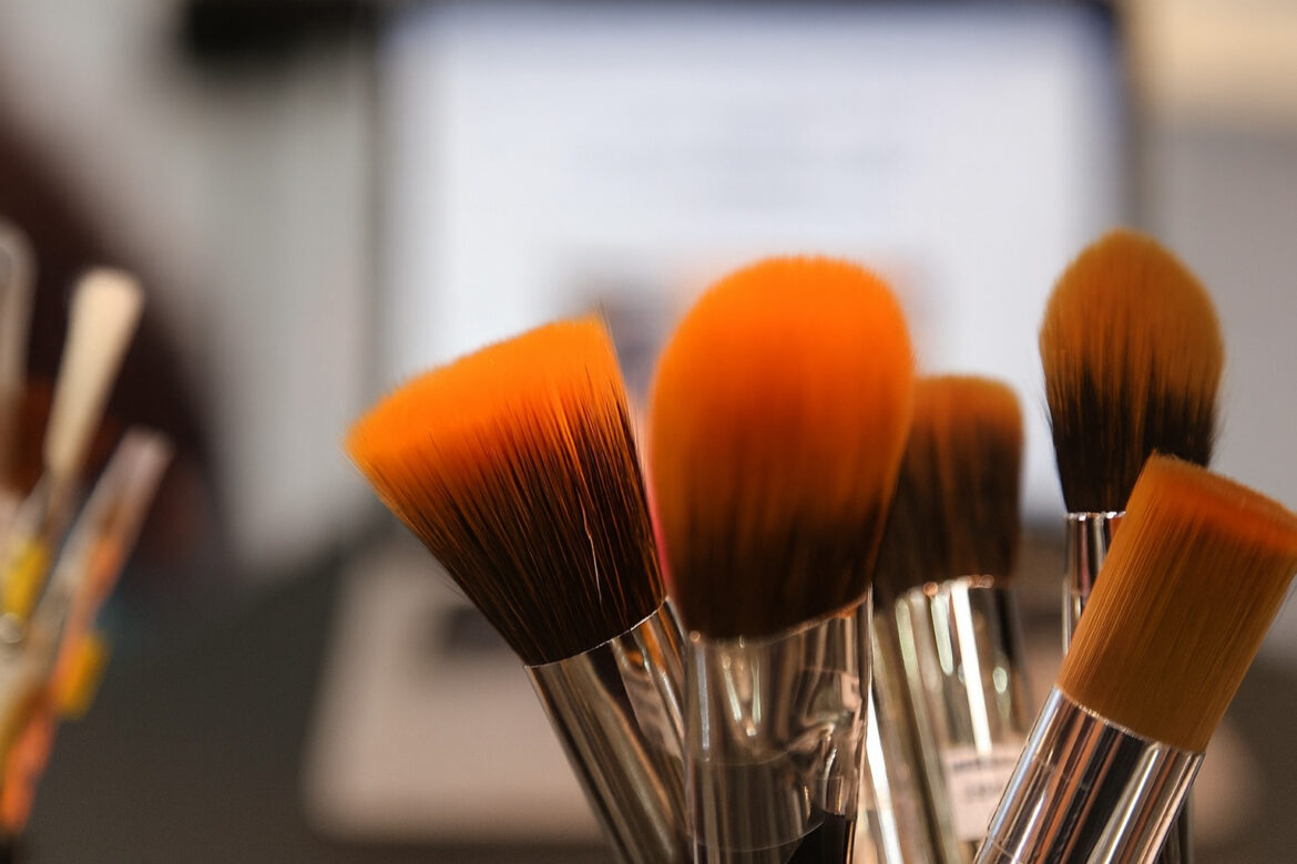 Nahaufnahme mehrerer Make-up-Pinsel mit orangefarbenen Borsten, im Hintergrund unscharf ein geöffneter Laptop.